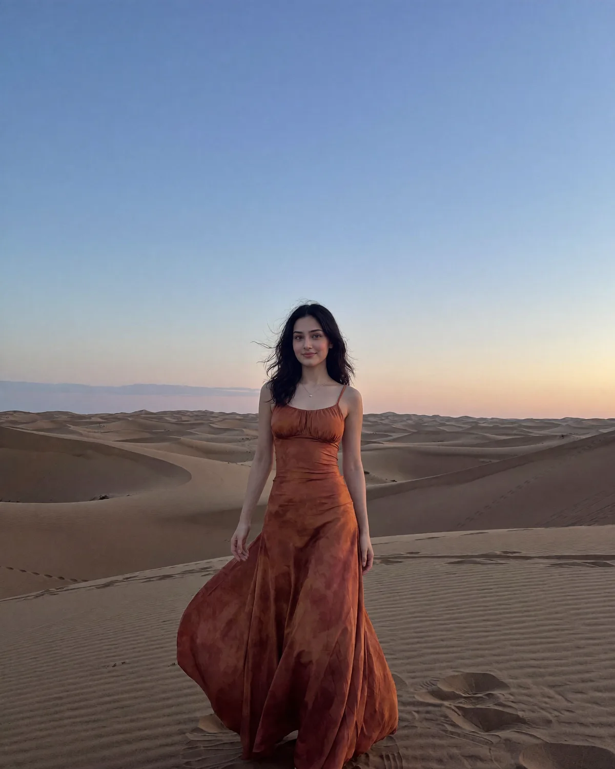A woman walking across desert dunes at blue hour in a long rust-coloured dress.