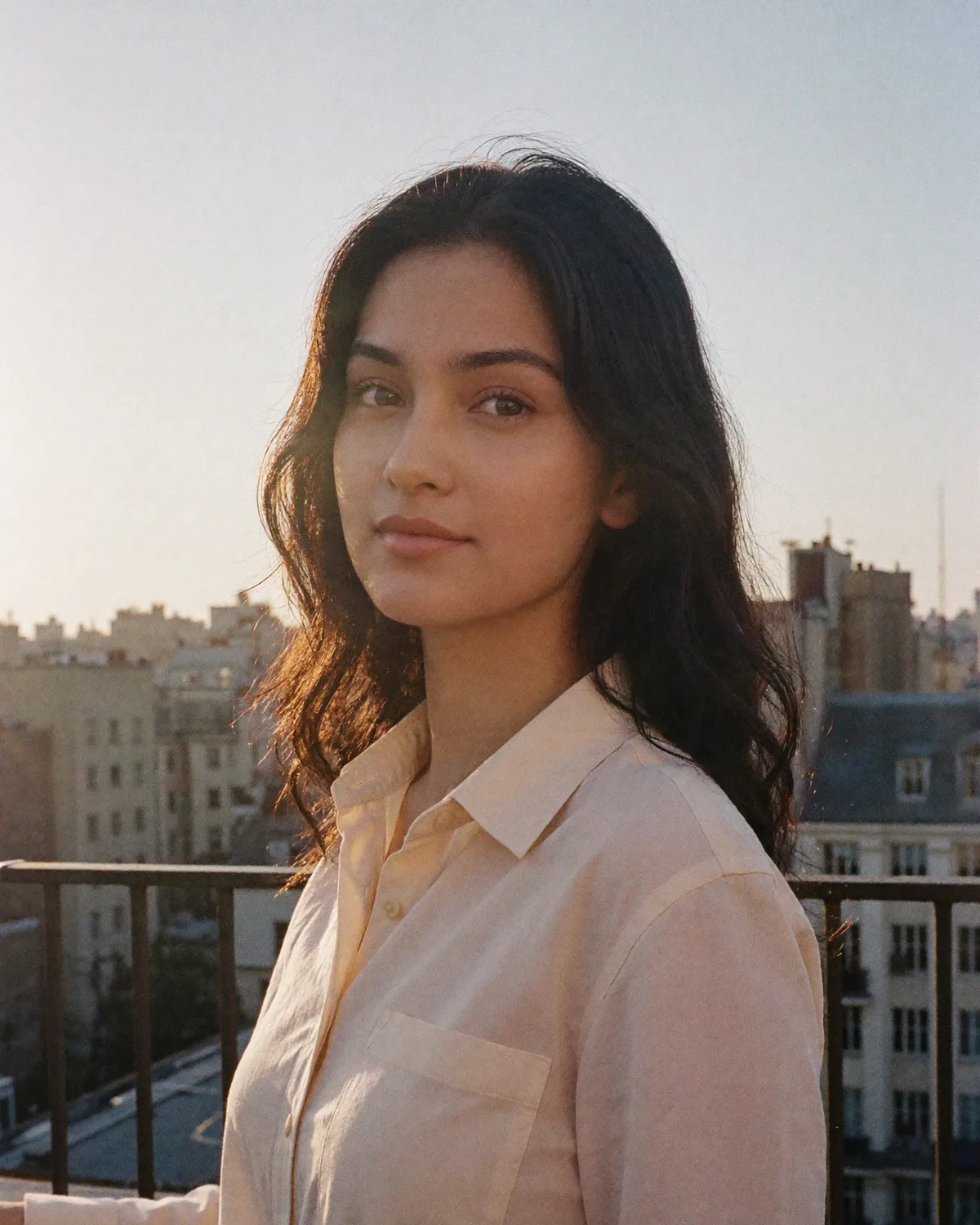 A woman on a city rooftop at golden hour, in a cream linen shirt, the skyline soft behind her.