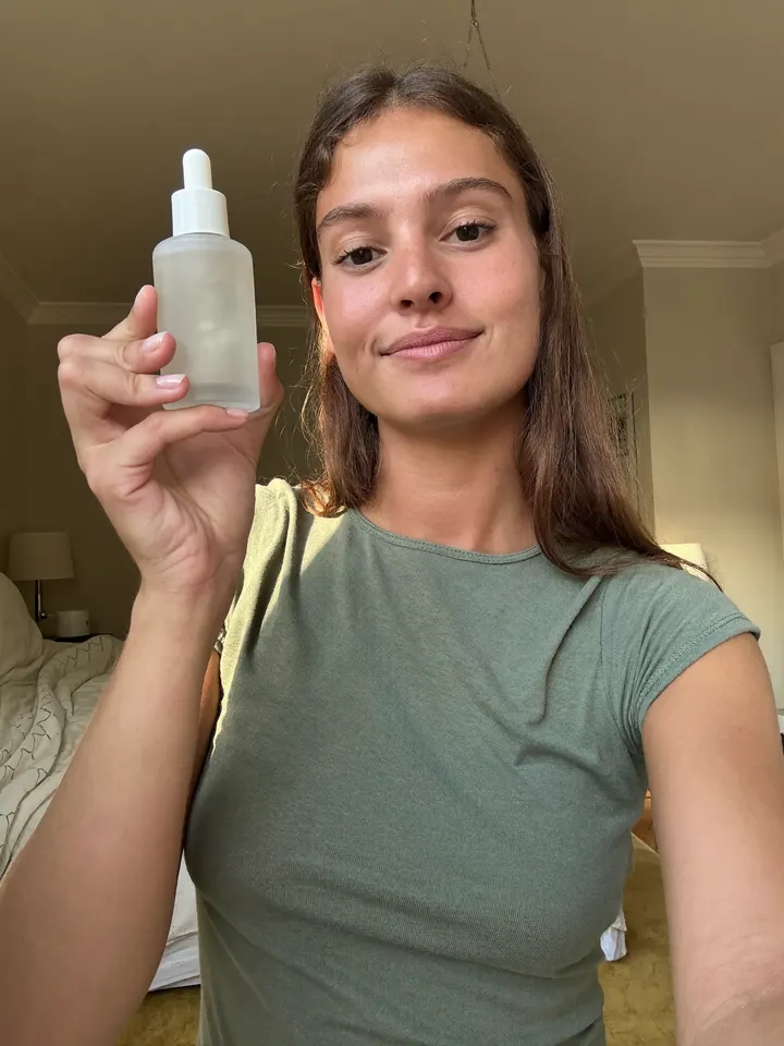 A woman filming a selfie video, holding up a bottle of face serum