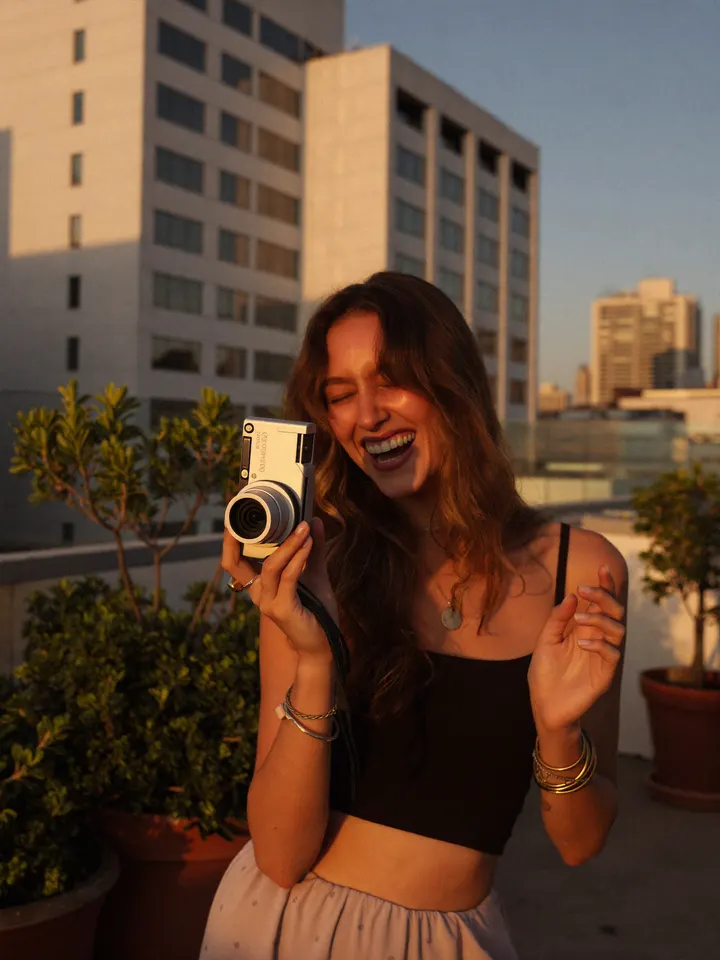 A woman laughing on a rooftop at golden hour, holding up a compact camera